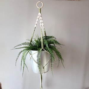 Puede incluir: Un colgador de plantas de macramé con un cubo de metal plateado lleno de helechos verdes. El macramé es blanco roto y verde oliva, con un anillo de madera en la parte superior para colgar. El colgador de plantas está suspendido contra una pared gris. La maceta mide unos 15 cm de diámetro.