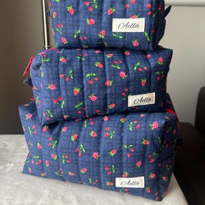 May include: Three quilted cosmetic bags stacked on top of each other. The bags are navy blue with a pattern of pink strawberries and green leaves. Each bag has a red zipper and a white label that says "Aella".