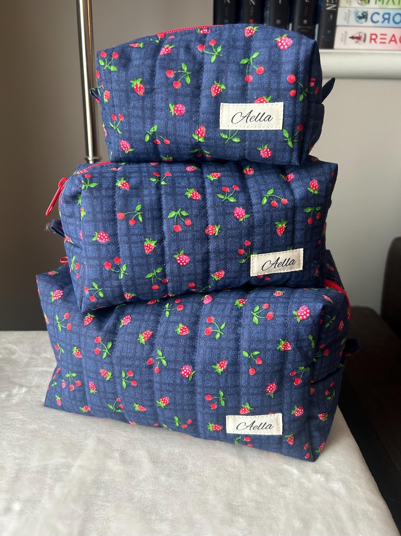 May include: Three quilted cosmetic bags stacked on top of each other. The bags are navy blue with a pattern of pink strawberries and green leaves. Each bag has a red zipper and a white label that says "Aella".