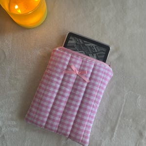 Może przedstawiać: Etui na czytnik e-booków w kratkę różową i białą z pikowanym wzorem i różową kokardką. Etui ma zamek błyskawiczny.