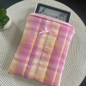Puede incluir: Funda para lector de libros electrónicos a cuadros rosas y amarillos con un pequeño lazo rosa. La funda está hecha de tela suave y tiene cierre de cremallera. El lector de libros electrónicos es parcialmente visible, mostrando el título "The Serpent".