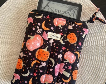 Könnte beinhalten: Schwarze, gesteppte E-Reader-Hülle mit einem skurrilen Muster aus Kürbissen, Pilzen, Monden und Sternen in Orange, Rosa und Weiß. Die Hülle hat einen schwarzen Reißverschluss und ein Etikett mit der Aufschrift "Aella". Der E-Reader zeigt "The Serpent".