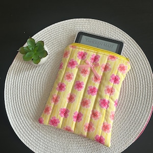 Könnte beinhalten: Eine gelb gesteppte Tablethülle mit einem rosa Blumenmuster und einer kleinen rosa Schleife. Die Hülle hat einen gelben Reißverschluss und enthält ein Tablet mit dem Text "TAHEREH MAFI". Die Hülle liegt auf einem weißen gewebten Platzset.