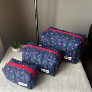 May include: Three navy blue quilted cosmetic bags with a red zipper and a floral pattern of pink flowers and red berries. Each bag has a white label with the name "Aella" in cursive.