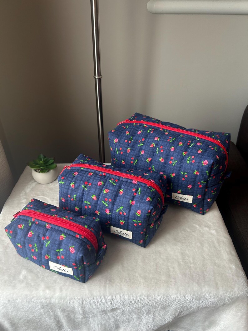 May include: Three navy blue quilted cosmetic bags with a red zipper and a floral pattern of pink flowers and red berries. Each bag has a white label with the name "Aella" in cursive.