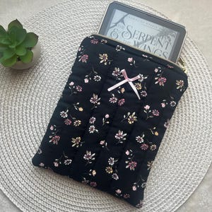 Puede incluir: Funda acolchada negra para lector de libros electrónicos con un estampado floral en rosa, amarillo y blanco. La funda tiene una cremallera dorada y un pequeño lazo rosa. Un lector de libros electrónicos es parcialmente visible.