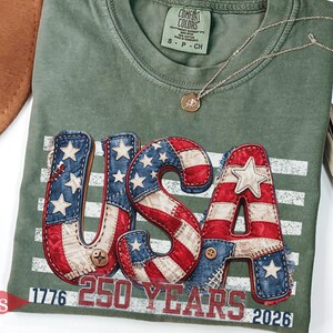 Puede incluir: Camiseta verde musgo con un diseño patriótico "USA" en rojo, blanco y azul, con estrellas y rayas. La camiseta incluye el texto "1776 250 YEARS 2026". Un collar dorado con un pequeño colgante descansa sobre la camiseta.