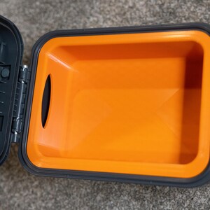 May include: A rectangular storage container with a dark gray exterior and a bright orange interior lining. The lid is hinged and open, revealing a smooth, hollow compartment with a single oval cutout on the side wall for easy access or handling.