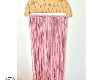 May include: A wooden half-circle wall hanging with a laser-etched design of flowers and butterflies. The hanging has a pink yarn fringe.
