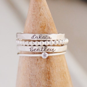 Puede incluir: Un conjunto de tres anillos de plata apilados en un soporte de anillo de madera. El anillo superior tiene el nombre "Dakota" grabado. El anillo del medio es un anillo de cuentas. El anillo inferior tiene el nombre "Bentley" grabado y una pequeña piedra transparente en el centro.