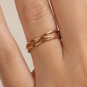 Op de afbeelding: Een goudkleurige ring met een delicaat ontwerp van meerdere strengen, waarbij dunne, gepolijste metalen banden elkaar kruisen in een doorlopend, vloeiend patroon. De ring heeft een glad, reflecterend oppervlak en een slank profiel rond de vinger.