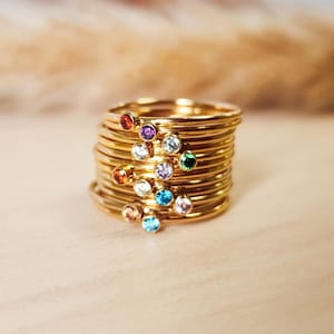 May include: A stack of gold-colored rings, each with a small, colorful gemstone. The gemstones are in various colors, including red, orange, yellow, green, blue, and purple. The rings are stacked on a light-colored wooden surface.