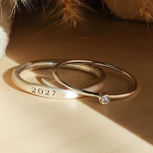 Puede incluir: Dos anillos de plata. Un anillo está grabado con el número "2027". El otro anillo es una banda fina con una pequeña gema transparente. Los anillos se exhiben sobre una superficie de color claro, con plantas secas en el fondo.