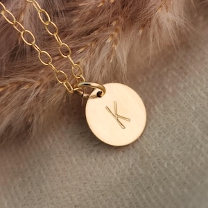 May include: A gold necklace with a fine chain and a circular pendant. The pendant is engraved with the letter "K". The necklace is set against a soft, neutral backdrop with dried plants, creating a minimalist aesthetic.