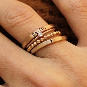May include: A set of four gold rings stacked on a finger. The top ring has the word "faith" engraved on it. The second ring has a small clear stone. The third ring is a beaded ring. The bottom ring has a small cross engraved on it.