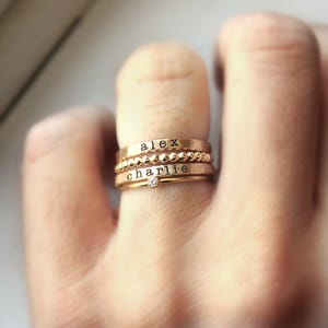 A hand wearing a stack of three gold-colored rings on a finger, engraved with the names alex and charlie, alongside a crystal solitaire ring.