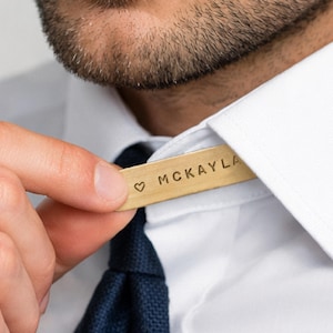 A man's hand adjusts a brass collar stay engraved "♡ MCKAYLA" in the collar of his white shirt, above a dark blue tie.