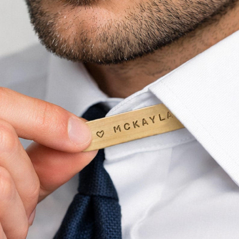 A man's hand adjusts a brass collar stay engraved "♡ MCKAYLA" in the collar of his white shirt, above a dark blue tie.