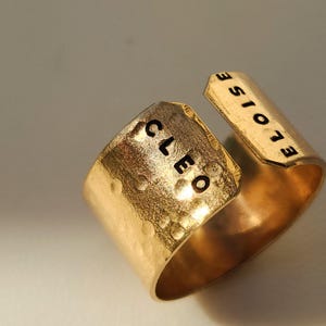 May include: A gold-colored ring with the name "CLEO" stamped on the front and "ELOISE" on the side. The ring has a hammered texture and a split design.