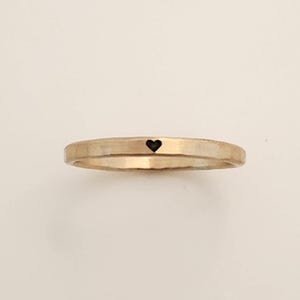 May include: A gold-coloured ring with a hammered texture and a small black heart. The ring is a simple band design, suitable for everyday wear. The heart is centered on the band.