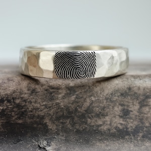 Puede incluir: Un anillo de plata con un acabado martillado y texturizado que presenta un patrón de huella dactilar grabado, oscuro y prominente en la superficie frontal. La banda tiene un brillo metálico pulido y un perfil redondeado con un diseño en espiral.