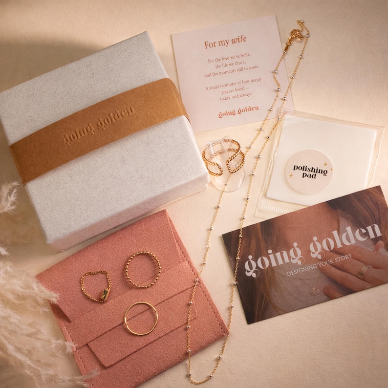 A white gift box with a brown band, a gold beaded necklace, two gold rings, a pink pouch, a polishing pad, and a "going golden" card are arranged on a surface.