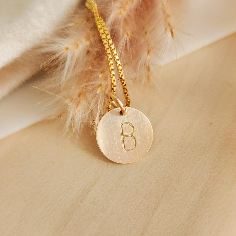 May include: A gold-coloured necklace with a circular pendant bearing the letter "B". The pendant is suspended from a fine chain. The background features soft, neutral tones and a hint of dried floral elements.