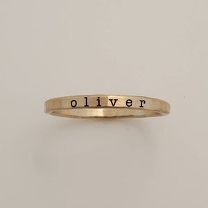 A close-up of a thin, hammered gold ring with the name 'oliver' engraved in lowercase black letters, set against a plain, off-white background.