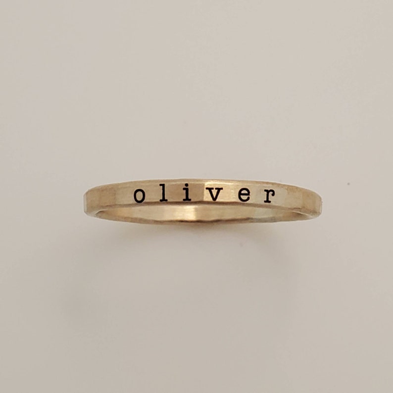 A close-up of a thin, hammered gold ring with the name 'oliver' engraved in lowercase black letters, set against a plain, off-white background.