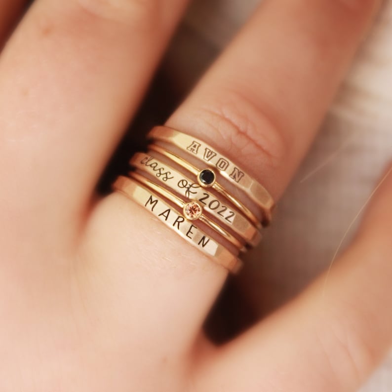 A five piece yellow gold class ring set. The set consists of three hand made, lightly hammered rings with the word 'Avon' engraved on one, '2022' engraved on one and the name 'Maren' engraved on one. All are engraved in black. The rings are separated by yellow gold stacking birthstone rings, one yellow and one black. The rings are on the hand of a woman wearing a white shirt.