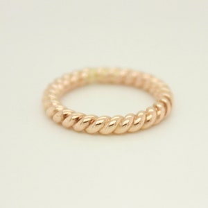 May include: A rose gold ring with a twisted rope design. The ring is a continuous band with a textured surface, reflecting light. The jewelry piece is set against a plain white background, highlighting its metallic sheen.