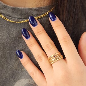 A hand with dark blue nails wearing a stack of three gold-colored name rings with a purple gemstone on a finger.