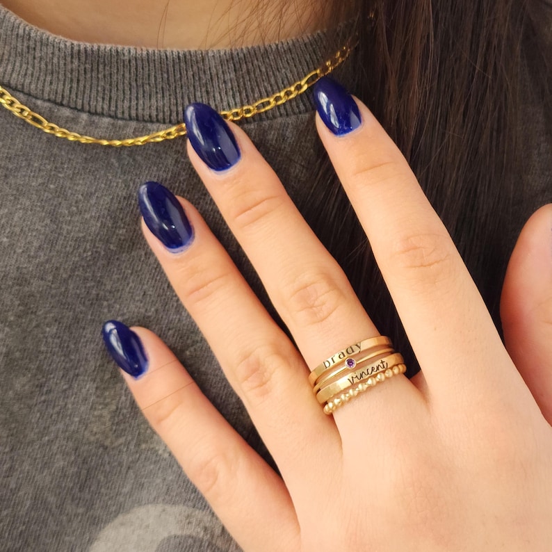 A hand with dark blue nails wearing a stack of three gold-colored name rings with a purple gemstone on a finger.