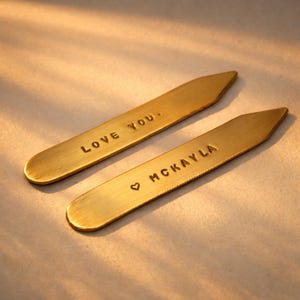 Two brass collar stays, one engraved "LOVE YOU." and the other "♡ MCKAYLA", are on a light surface with soft shadows.