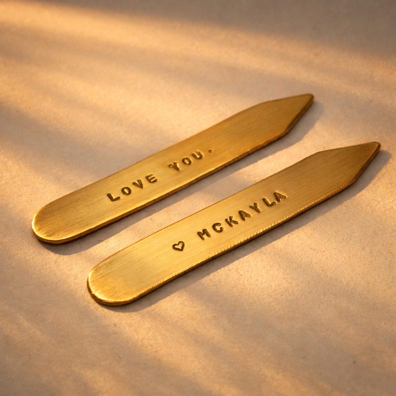 Two brass collar stays, one engraved "LOVE YOU." and the other "♡ MCKAYLA", are on a light surface with soft shadows.
