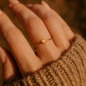 May include: A delicate gold ring featuring a small heart-shaped charm. The ring is thin and simple, with a polished finish. The heart charm has a textured surface. The ring is shown on a finger, against a background of a brown sweater.