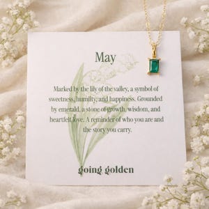 May include: A gold-colored necklace with a rectangular emerald pendant hangs above a card. The card reads "May" and includes text about the lily of the valley and emeralds. The words "going golden" are at the bottom.