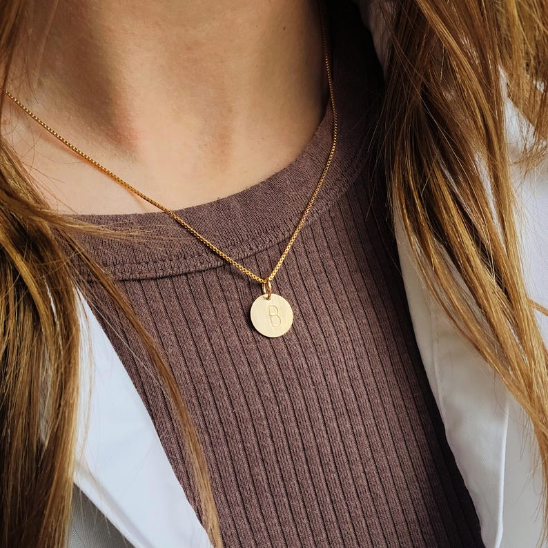 May include: A gold necklace with a circular pendant. The pendant has the letter "B" in the centre. The necklace is worn against a brown ribbed top and a white shirt.