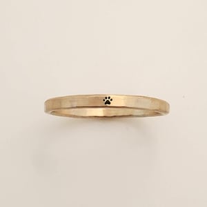 Puede incluir: Un anillo de color dorado con un pequeño diseño de huella de pata negra. El anillo tiene un diseño de banda simple y una superficie ligeramente texturizada. Este anillo es un accesorio con temática de mascotas.