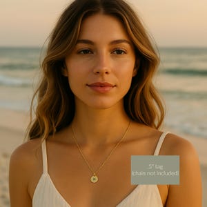 May include: A gold-colored necklace with a small, round pendant featuring the letter "H". The pendant is approximately 0.5 inches in diameter. The necklace is displayed on a person wearing a white top, with a beach and ocean background.