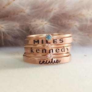 Puede incluir: Una pila de anillos de oro rosa, cada uno grabado con un nombre: Miles, Kennedy y Cecilia. Cada anillo tiene una pequeña piedra preciosa de colores. Los anillos están apilados uno encima del otro.