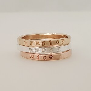 Puede incluir: Tres anillos apilables en oro, plata y oro rosa. Cada anillo tiene una textura martillada y letras estampadas que deletrean los nombres "bradley", "brody" y "nico". Los anillos se muestran sobre una superficie blanca.