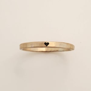 May include: A gold-colored ring with a hammered texture and a small black heart. The ring is a simple band design, suitable for everyday wear. The heart is centered on the band.
