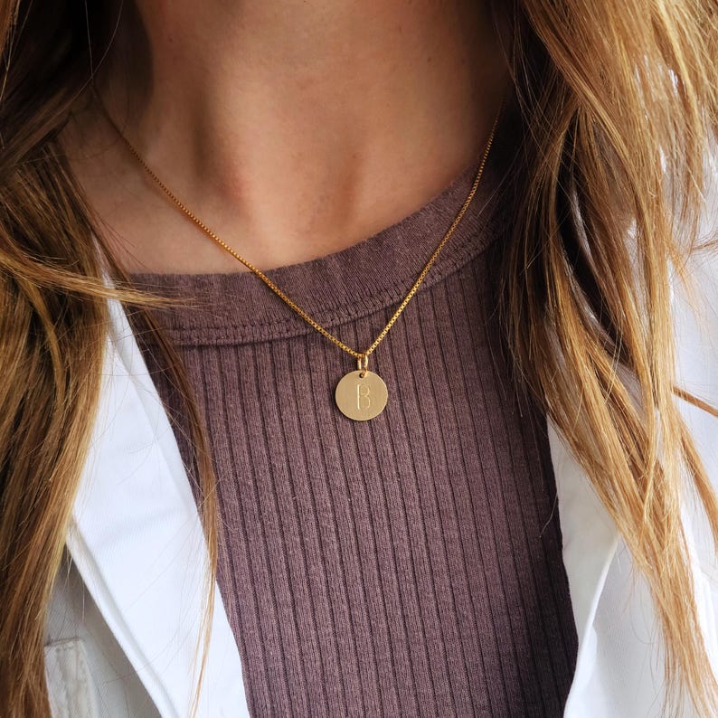 May include: Gold-coloured necklace with a circular pendant. The pendant features a single letter "B" in a simple font. The necklace is worn against a brown ribbed top and a white jacket.