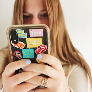 A five piece yellow gold class ring set. The set consists of three hand made, lightly hammered rings with the word 'Avon' engraved on one, '2022' engraved on one and the name 'Maren' engraved on one. All are engraved in black. The rings are separated by yellow gold stacking birthstone rings, one yellow and one black. The rings are on the hand of a woman wearing a white shirt, holding a phone with bright colored stickers on it.