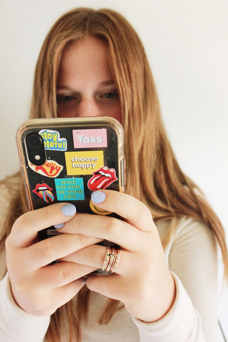 A five piece yellow gold class ring set. The set consists of three hand made, lightly hammered rings with the word 'Avon' engraved on one, '2022' engraved on one and the name 'Maren' engraved on one. All are engraved in black. The rings are separated by yellow gold stacking birthstone rings, one yellow and one black. The rings are on the hand of a woman wearing a white shirt, holding a phone with bright colored stickers on it.