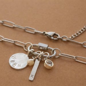A silver paperclip chain necklace with a carabiner clasp holds four charms: a round disc with "OWEN" stamped on it, a small white pearl, a silver bar with "K" stamped on it, and a round clear gemstone in a bezel setting, with the chain extending