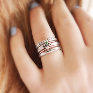 May include: A collection of silver rings worn on a finger. One ring is inscribed with "ZIONSVILLE" and features a green gemstone. Another ring displays the number "2024" and a clear gem. The bottom ring is engraved with the name "maddy".