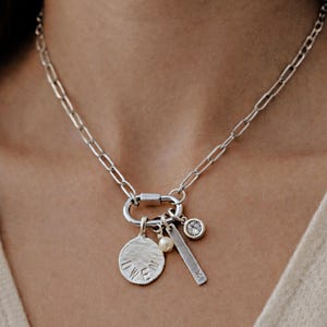 Pode incluir: Colar de prata com um grande fecho de mosquetão e vários pingentes. Os pingentes incluem um disco texturizado com o nome "OWEN", uma pequena pérola, uma barra retangular com a letra "K" e um pingente redondo com uma pedra transparente.