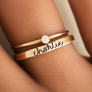 Peut inclure: Deux bagues dorées empilées. La bague supérieure est un anneau fin avec une petite pierre ronde transparente sertie clos. La bague inférieure est un anneau plat gravé du nom charlie dans une police d'écriture cursive de couleur noire.